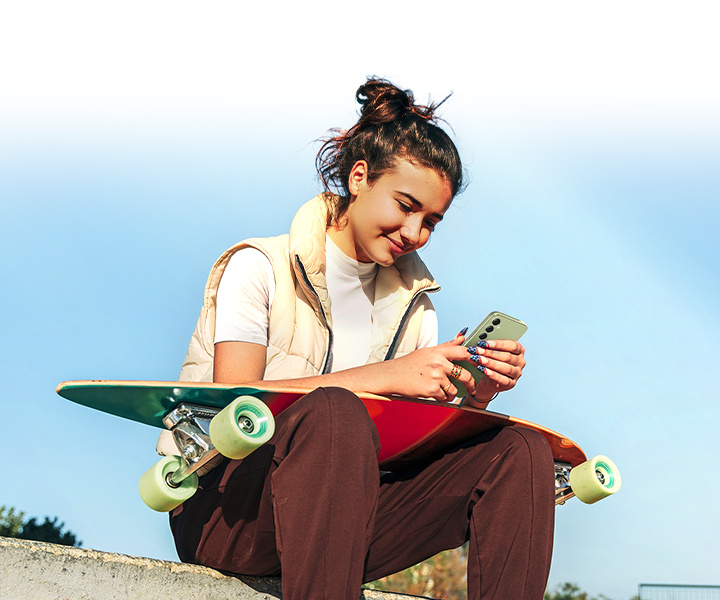A woman is looking at Galaxy A54 5G under bright sunlight, with a skateboard on her lap.