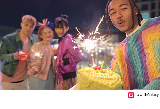 Three friends are on top of a bridge at night, celebrating with sparklers. When another friend enters the frame from the right, holding a cake, the Auto-framing feature automatically adjusts the frame to include all of them as well as the focus on the friend holding the cake, who is standing closer to in the frame. Camera icon and #WithGalaxy text are shown.