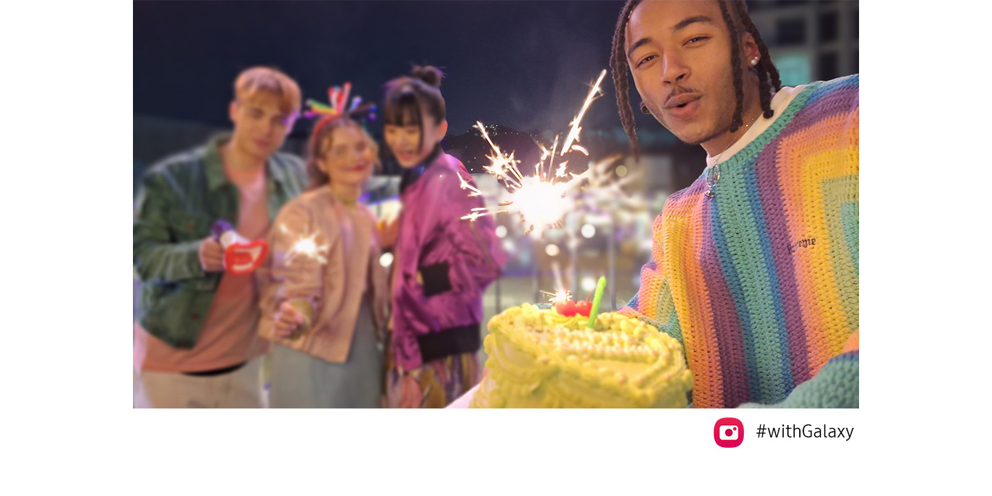 Three friends are on top of a bridge at night, celebrating with sparklers. When another friend enters the frame from the right, holding a cake, the Auto-framing feature automatically adjusts the frame to include all of them as well as the focus on the friend holding the cake, who is standing closer to in the frame. Camera icon and #WithGalaxy text are shown.