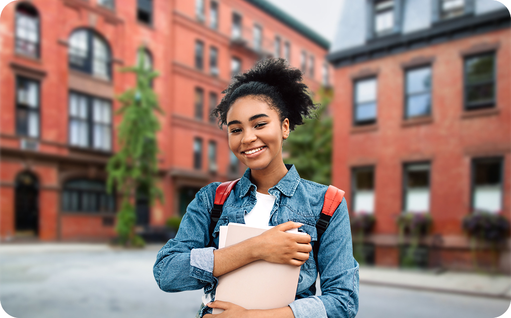 A female student, wearing a backpack and clutching onto a few books, is smiling. With the Portrait On icon above activated, she is in focus, and red brick-stoned buildings in the background are gently blurred.