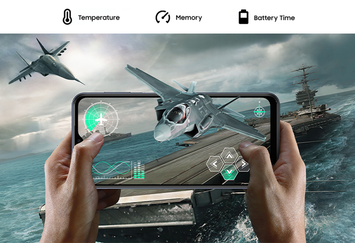 Two hands holding a Galaxy M33 5G device in horizontally, playing a fighter plane fps game. The background shows the in-game naval battlefield atmosphere with a large aircraft carrier extended to outside the screen. From inside the screen, a fighter plane is slightly protruding outside the screen. Above, text reads Temperature, Memory and Battery Time.