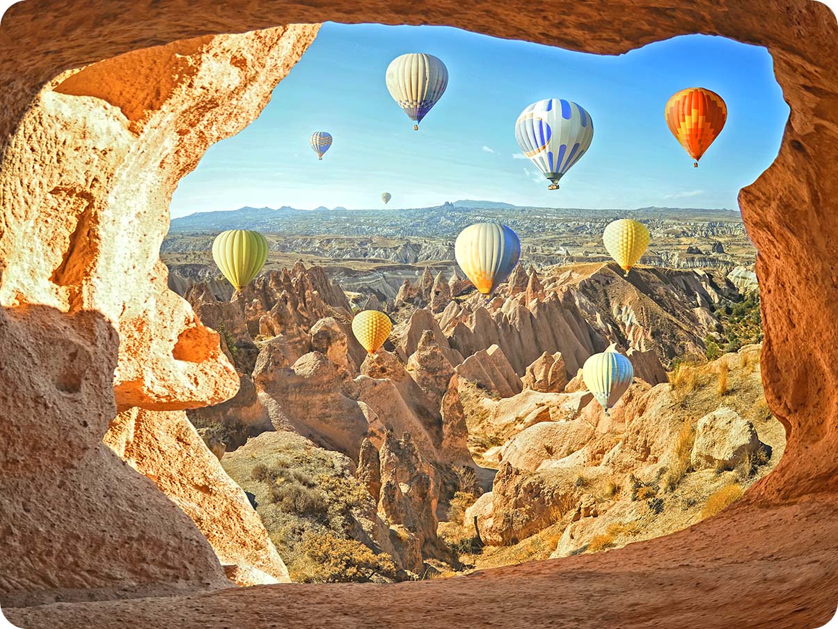 Photo taken with Ultra Wide Camera shows colorful air balloons in the sky and a vast landscape in the background. With the Ultra-wide angle icon above activated,  the edges of the shot include the cave where the shot was taken.