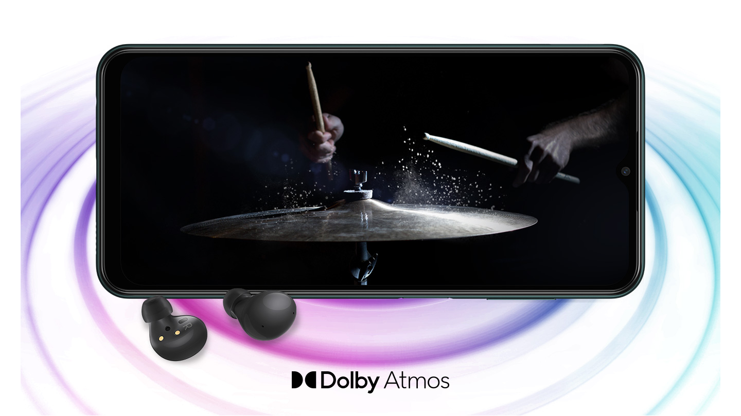 Galaxy M13 in landscape mode and an image with a person playing drums in the black background onscreen. A pair of black Galaxy Buds2 are placed in front of the device. On the bottom is a logo for Dolby Atmos.