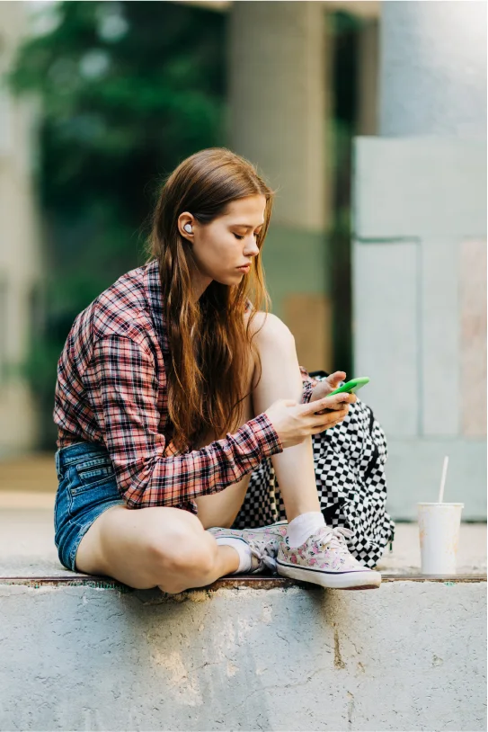 Young woman wearing JBL true wireless earbuds while using her smartphone outdoors