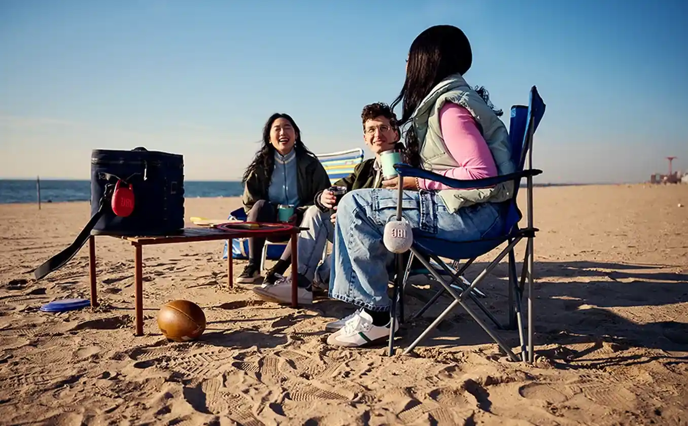 Group of friends at beach with JBL Clip 5 speakers clipped to chairs and bags enjoying multi-speaker audio