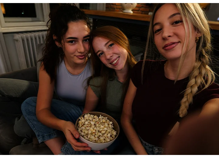 Three girls on a sofa at home in a low-lit environment, taking a photo together. One of the girls is taking the photo using Galaxy A37 5G with Nightography for bright, vibrant photo details.