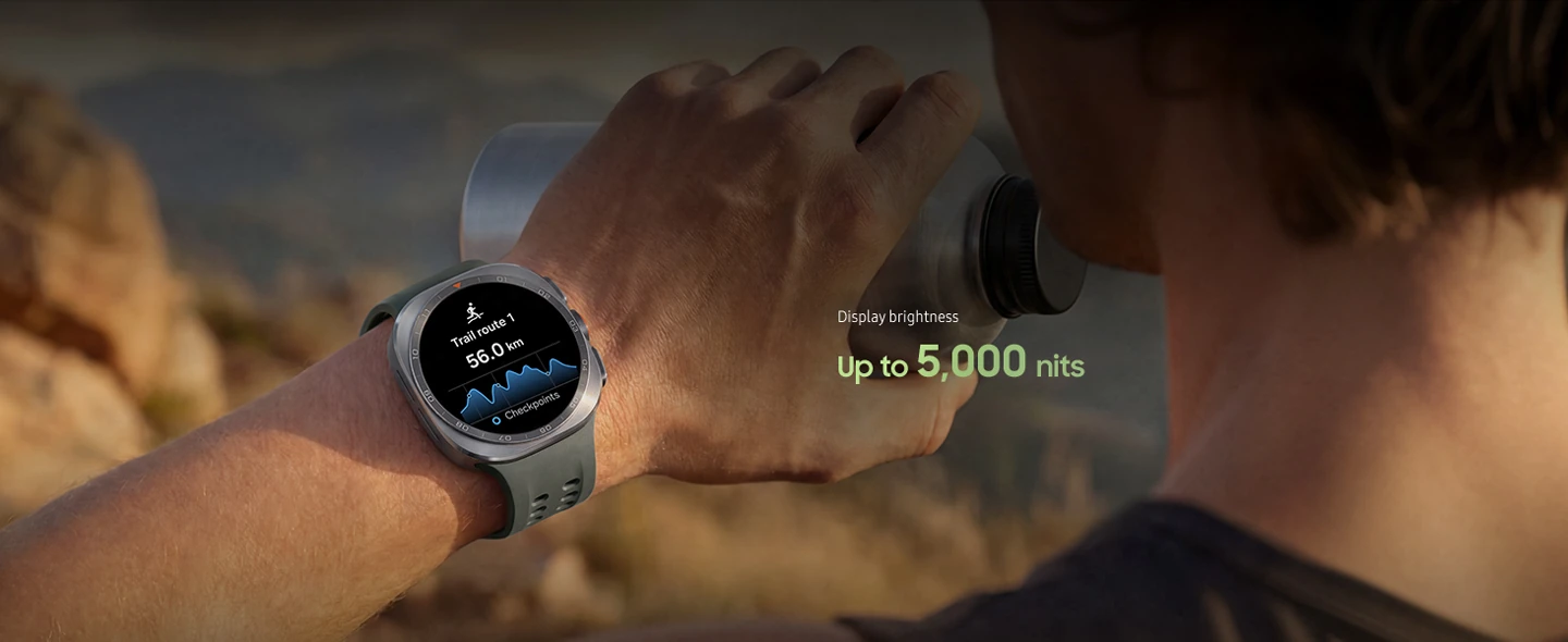 A man on a mountain trail with rocky terrain is holding a water tumbler in his hand while wearing a Galaxy Watch Ultra2 in Titanium Silver with a Marine Band in Olive, displaying a trail route with a distance, ideal for reliable guidance in challenging outdoor conditions during trail running and climbing.