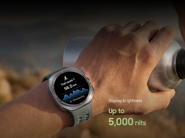 A man on a mountain trail with rocky terrain is holding a water tumbler in his hand while wearing a Galaxy Watch Ultra2 in Titanium Silver with a Marine Band in Olive, displaying a trail route with a distance, ideal for reliable guidance in challenging outdoor conditions during trail running and climbing.